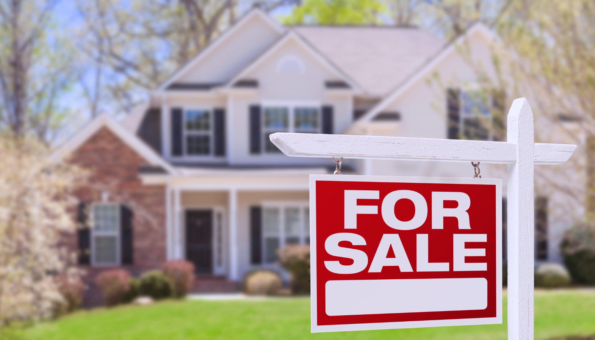 A traditional house in the background with a focus on a For Sale Sign